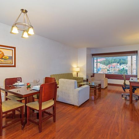 โรงแรมเตเกนดามา โบโกตา ภายนอก รูปภาพ The image shows a modern hotel room or living area. It features a cozy arrangement with a dining table and four chairs, a comfortable sofa, and a couple of armchairs. A large window provides a view of the outside, which includes greenery or possibly 