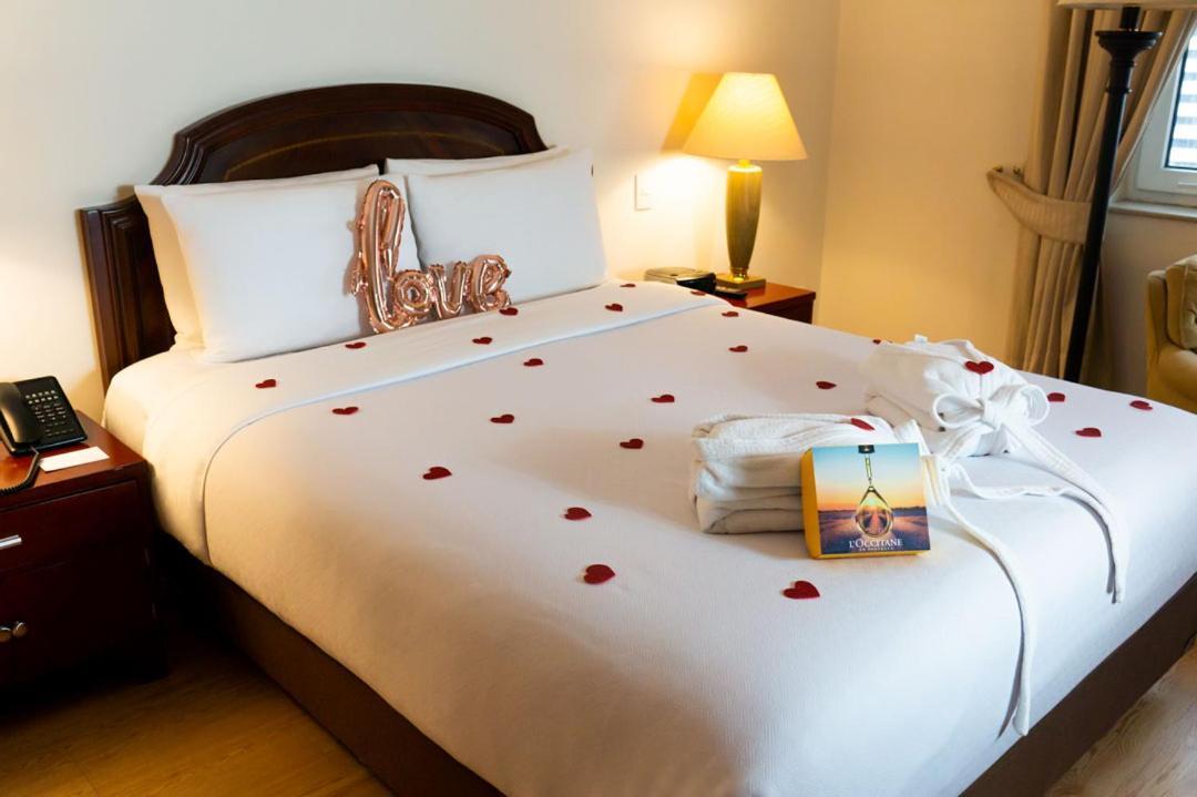 โรงแรมเตเกนดามา โบโกตา ภายนอก รูปภาพ The photo shows a neatly arranged bed in a hotel room. The bed has white linens and is adorned with rose petals scattered across the top. There is a decorative pillow that says "love" in a metallic finish. Next to the bed, there is a lamp providing s
