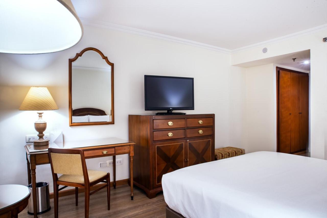 โรงแรมเตเกนดามา โบโกตา ภายนอก รูปภาพ The photo shows a hotel room featuring a bed with white linens, a wooden dresser with multiple drawers and a flat-screen television mounted on the wall above it. There is a mirror with a decorative frame hanging on the wall, and a lamp on a small sid