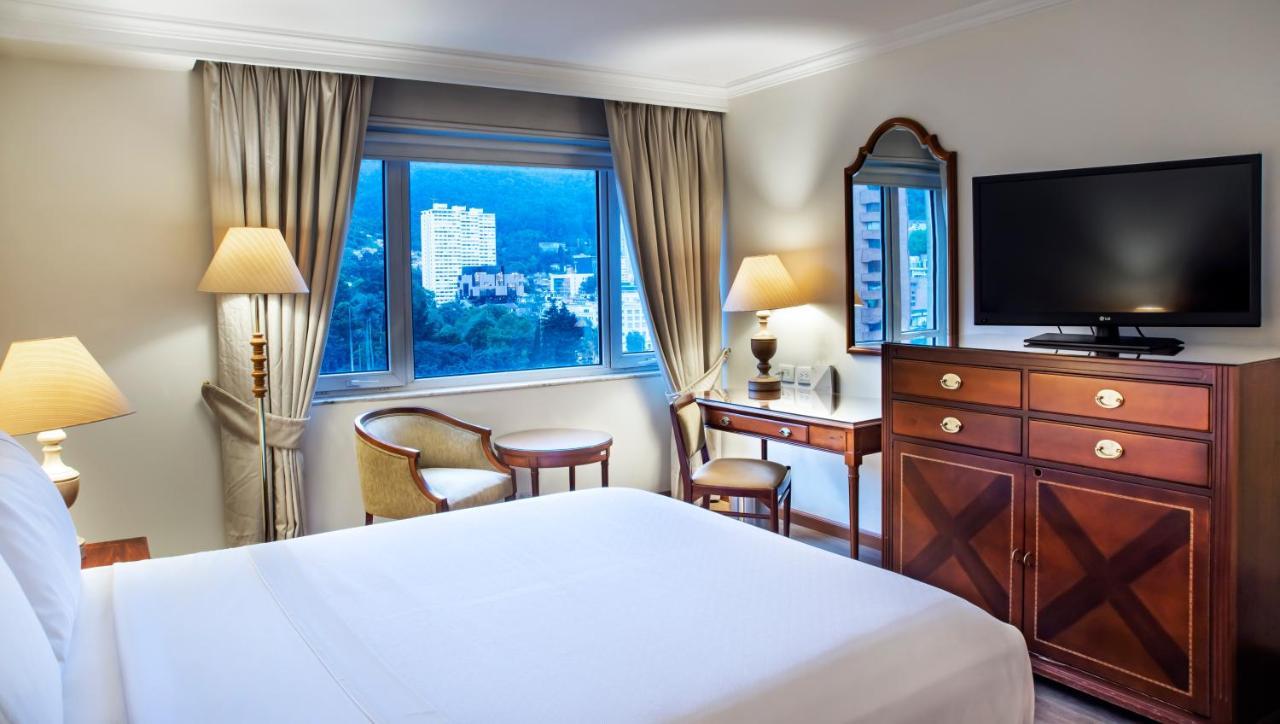 โรงแรมเตเกนดามา โบโกตา ภายนอก รูปภาพ The photo shows a well-lit hotel room featuring a large bed with a white bedspread. There are two bedside lamps on either side of the bed, providing soft lighting. A window with curtains allows natural light to enter the room, revealing a view of a c