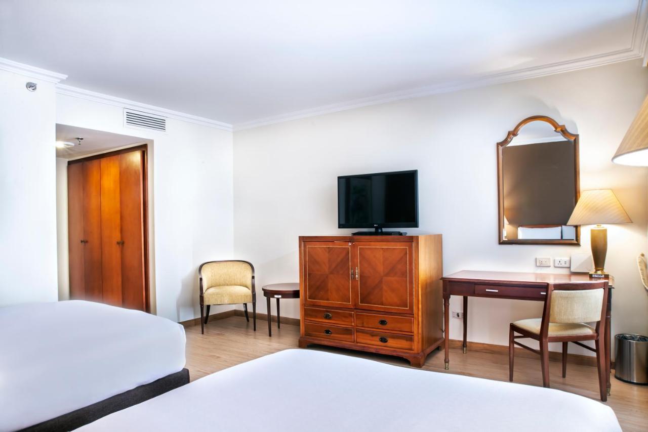 โรงแรมเตเกนดามา โบโกตา ภายนอก รูปภาพ The photo shows a well-furnished hotel room featuring two beds with white bedding. There is a wooden dresser or cabinet in the center, which likely contains storage space. A flat-screen TV is mounted on the wall above the dresser. To the right, there