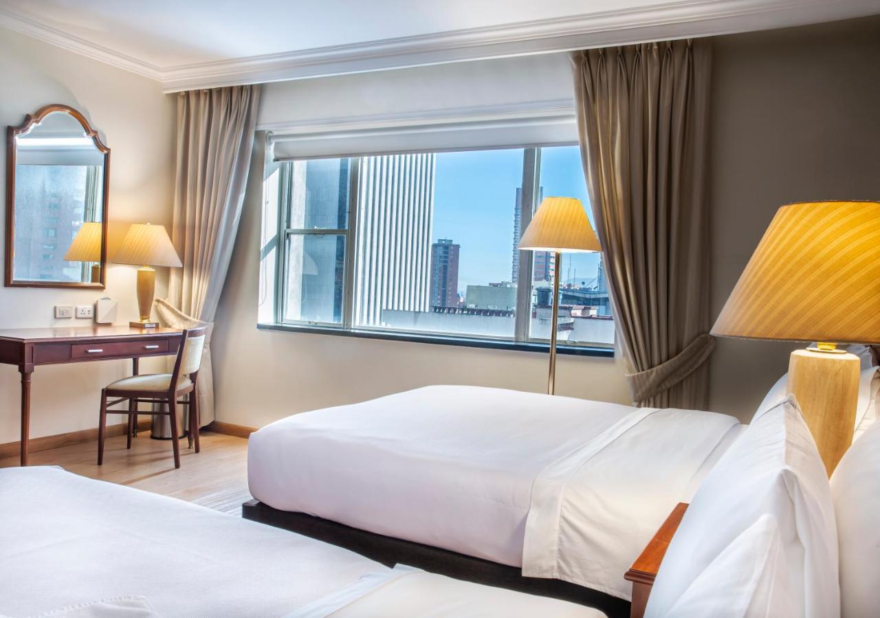 โรงแรมเตเกนดามา โบโกตา ภายนอก รูปภาพ The photo shows a hotel room featuring two beds with white linens. There is a large window allowing natural light to fill the space, with a view of a cityscape visible outside. The room is decorated with light-colored curtains, and there are two lamp