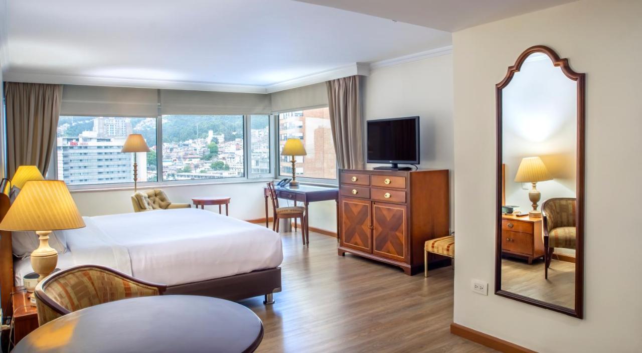 โรงแรมเตเกนดามา โบโกตา ภายนอก รูปภาพ The image shows a hotel room featuring a large window with natural light illuminating the space. There are two beds with white linens, a wooden dresser positioned against the wall, and a television mounted on a cabinet. A small desk and chair are in 