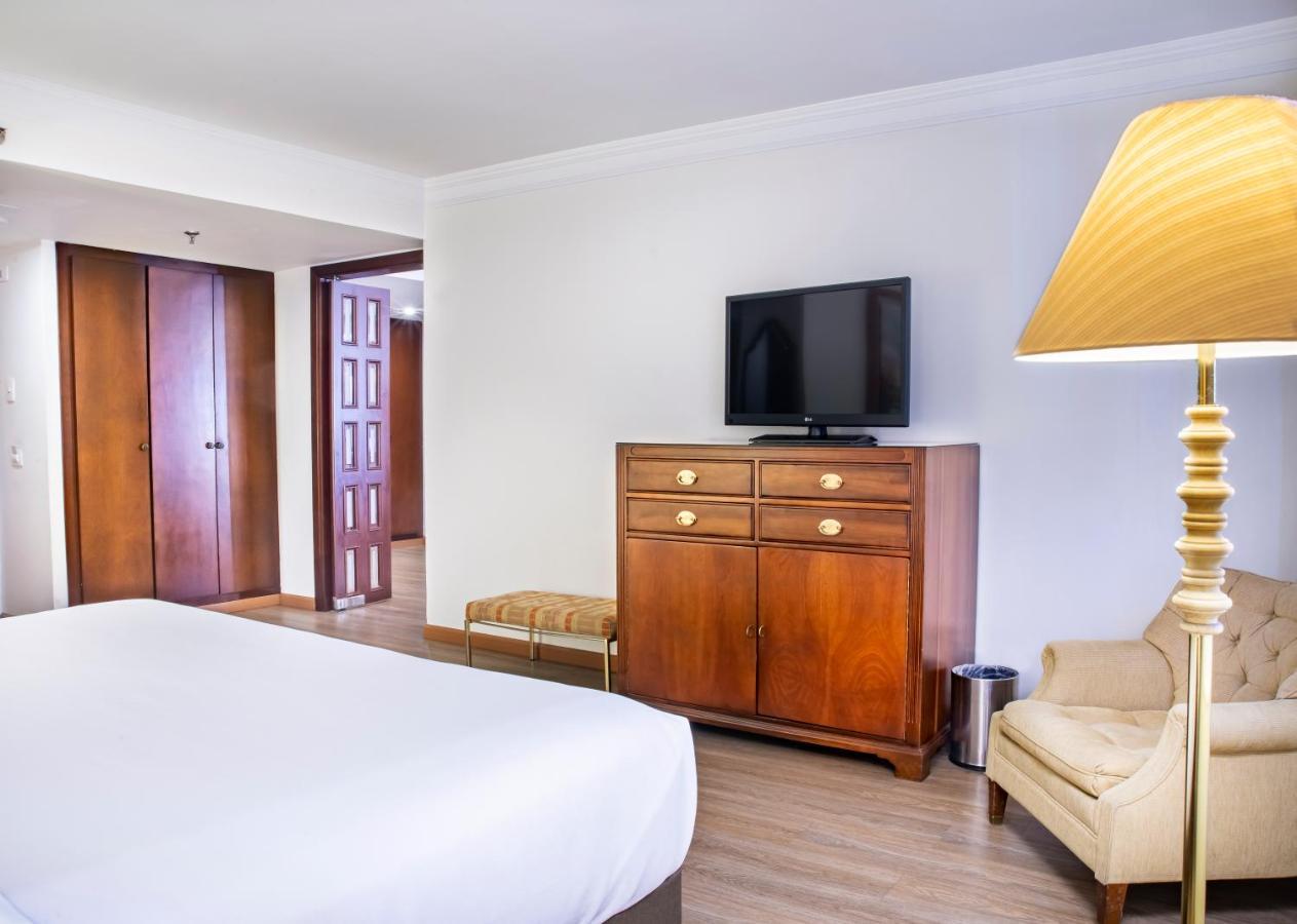 โรงแรมเตเกนดามา โบโกตา ภายนอก รูปภาพ The photo shows a simple hotel room with a clean and modern design. In the foreground, there is a bed with a white bedspread. To the right, there is a beige armchair and a tall lamp providing light. Against the wall, there is a wooden dresser with se
