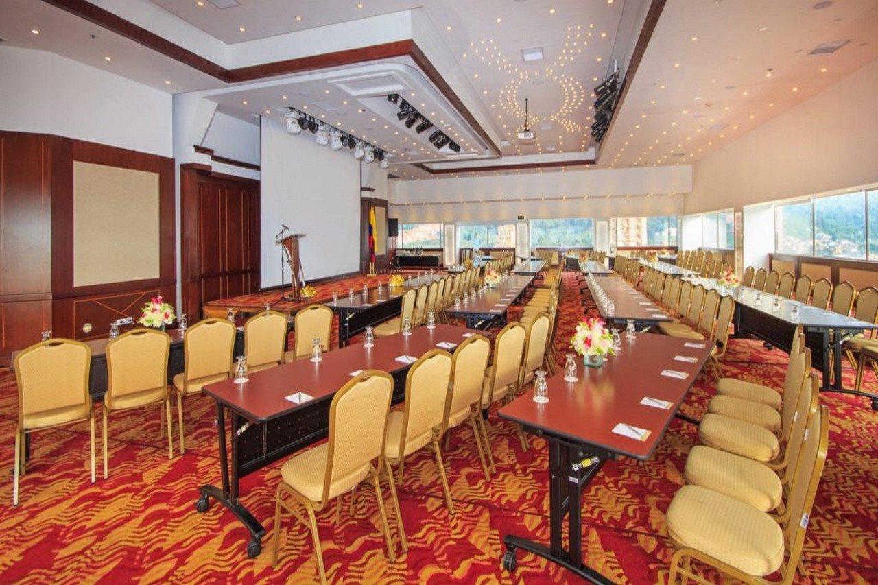 โรงแรมเตเกนดามา โบโกตา ภายนอก รูปภาพ The photo shows a spacious conference room set up for an event. There are several rows of tables arranged in a classroom-style layout, each accompanied by beige chairs. The floors are covered with a colorful, patterned carpet. At the front of the roo