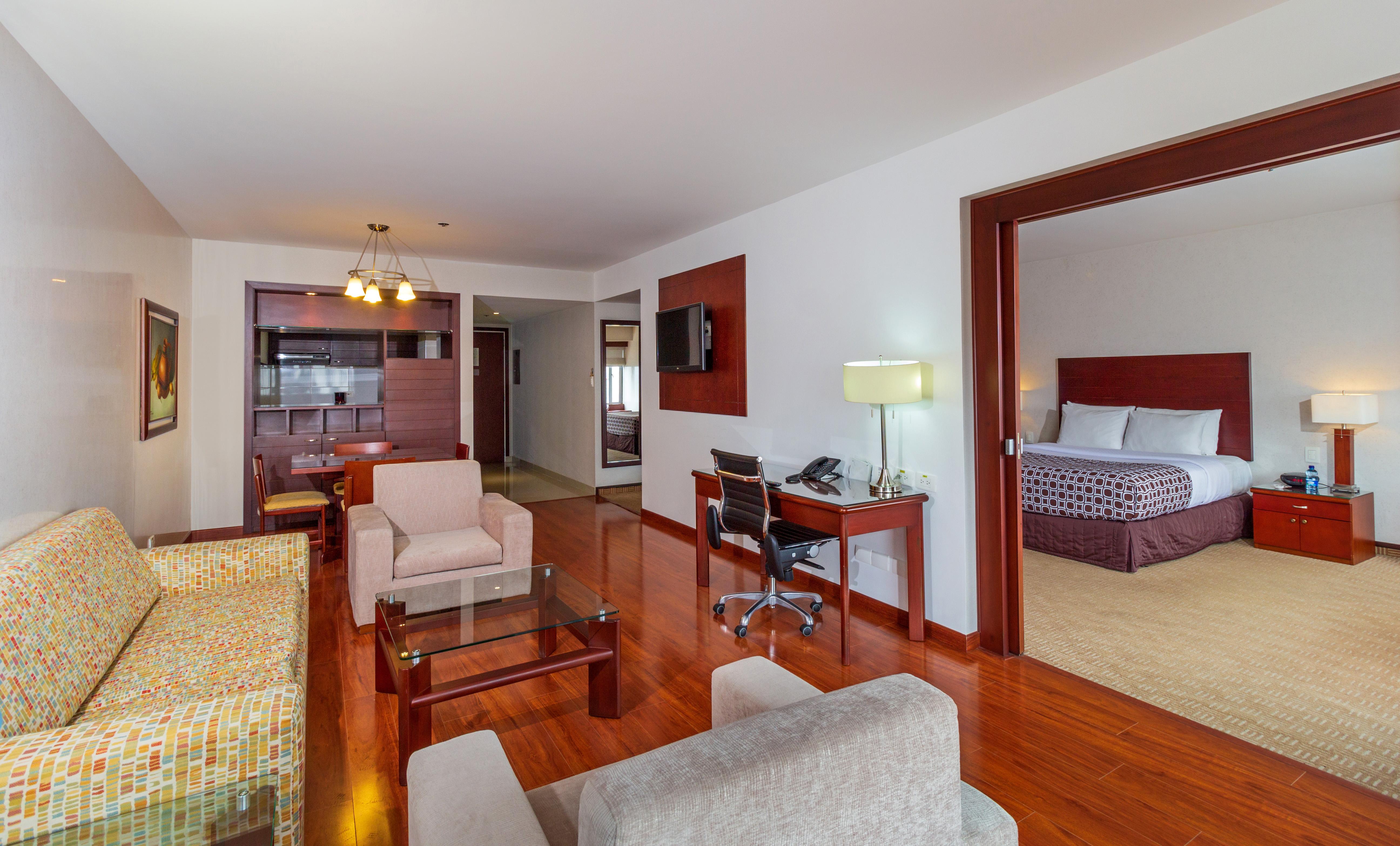 โรงแรมเตเกนดามา โบโกตา ภายนอก รูปภาพ The photo shows a hotel suite or a spacious living area. The room features hardwood floors and a layout that includes a seating area with two armchairs and a small table, along with a colorful sofa. There's a work desk with a chair, a television moun