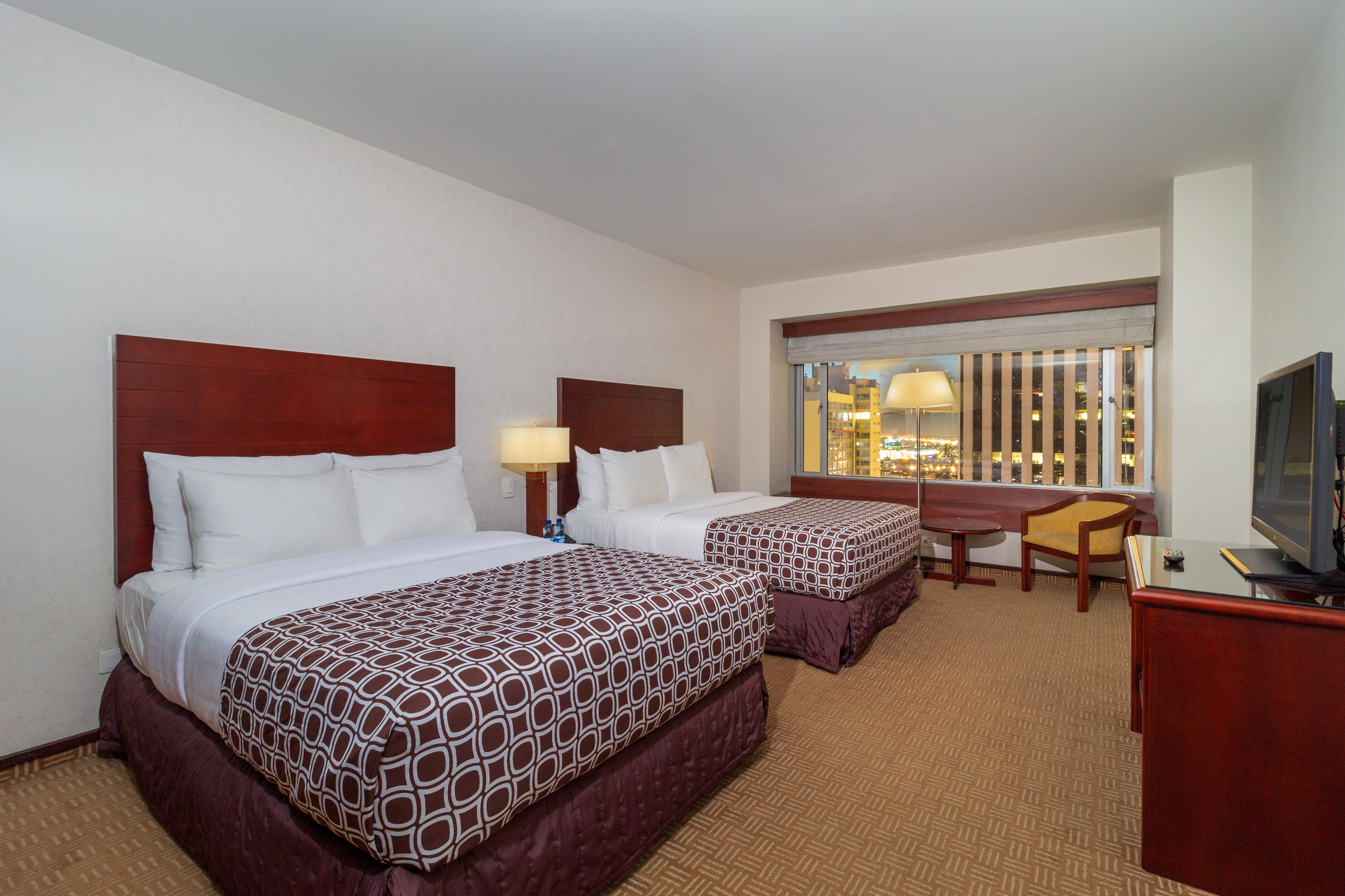 โรงแรมเตเกนดามา โบโกตา ภายนอก รูปภาพ The photo shows a hotel room featuring two beds with white linens and patterned bedspreads in dark tones. There is a bedside lamp next to one of the beds. A window is visible, providing light to the room, and it appears to offer a view outside. Addit