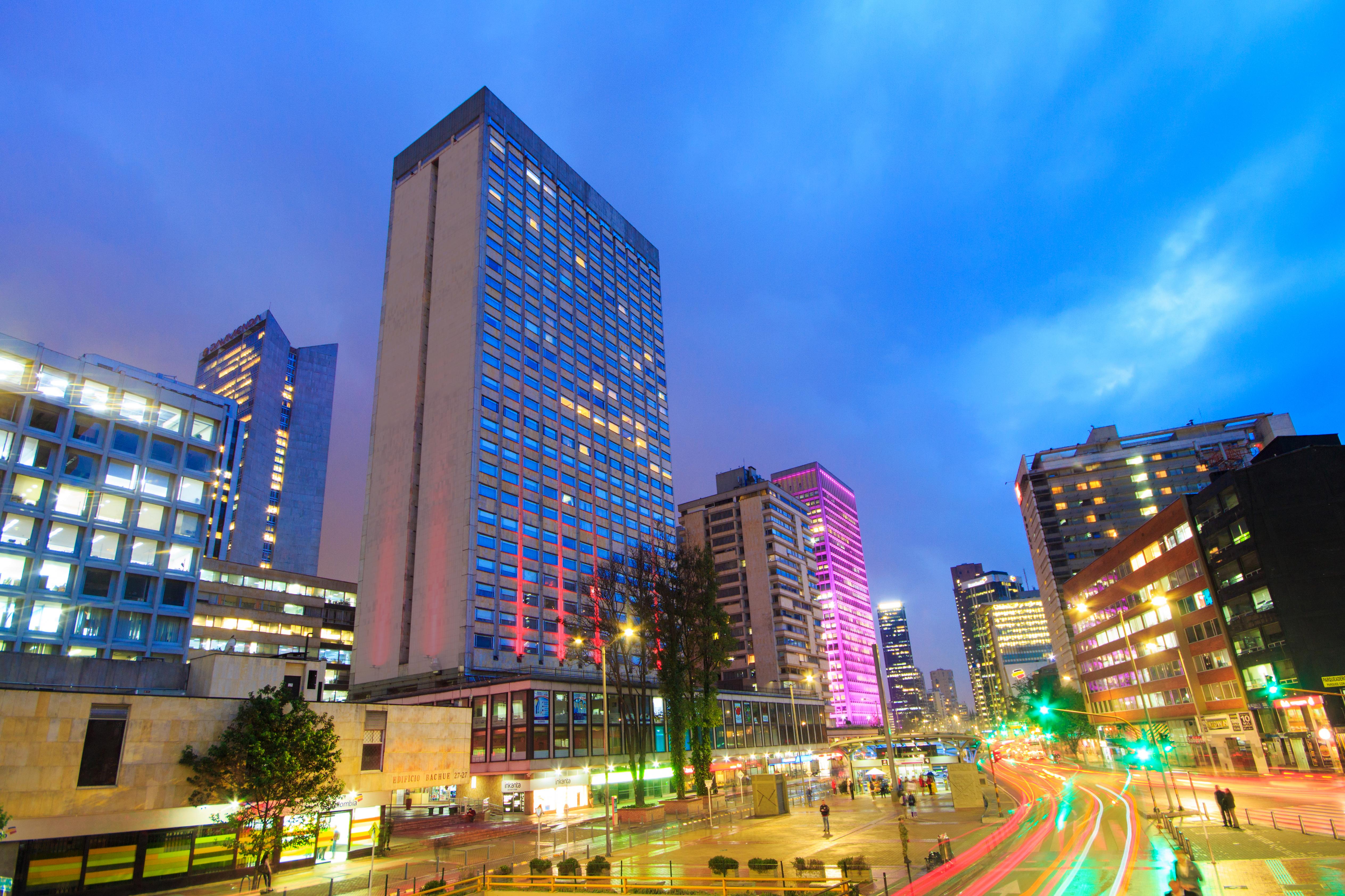 โรงแรมเตเกนดามา โบโกตา ภายนอก รูปภาพ The photo shows a vibrant urban landscape during dusk. Tall buildings are lit up with colorful lights, particularly in pink and blue hues. The sky is a gradient of colors, indicating twilight. There are trees and greenery along the streets, which app