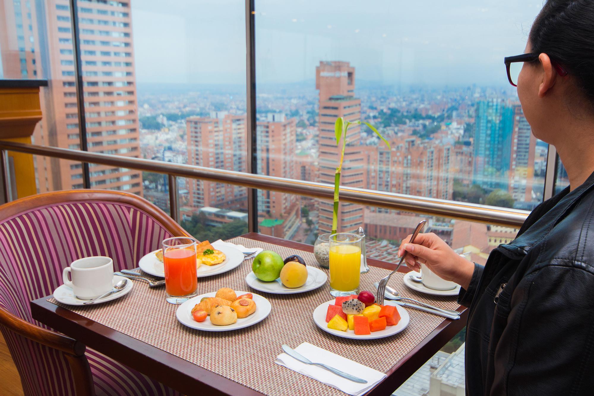โรงแรมเตเกนดามา โบโกตา ภายนอก รูปภาพ The photo depicts a breakfast scene at a table with a panoramic view of a city. The table is set with several plates: one with a colorful assortment of fresh fruit, another with small pastries or donuts, and a third with a beverage. There are two dri
