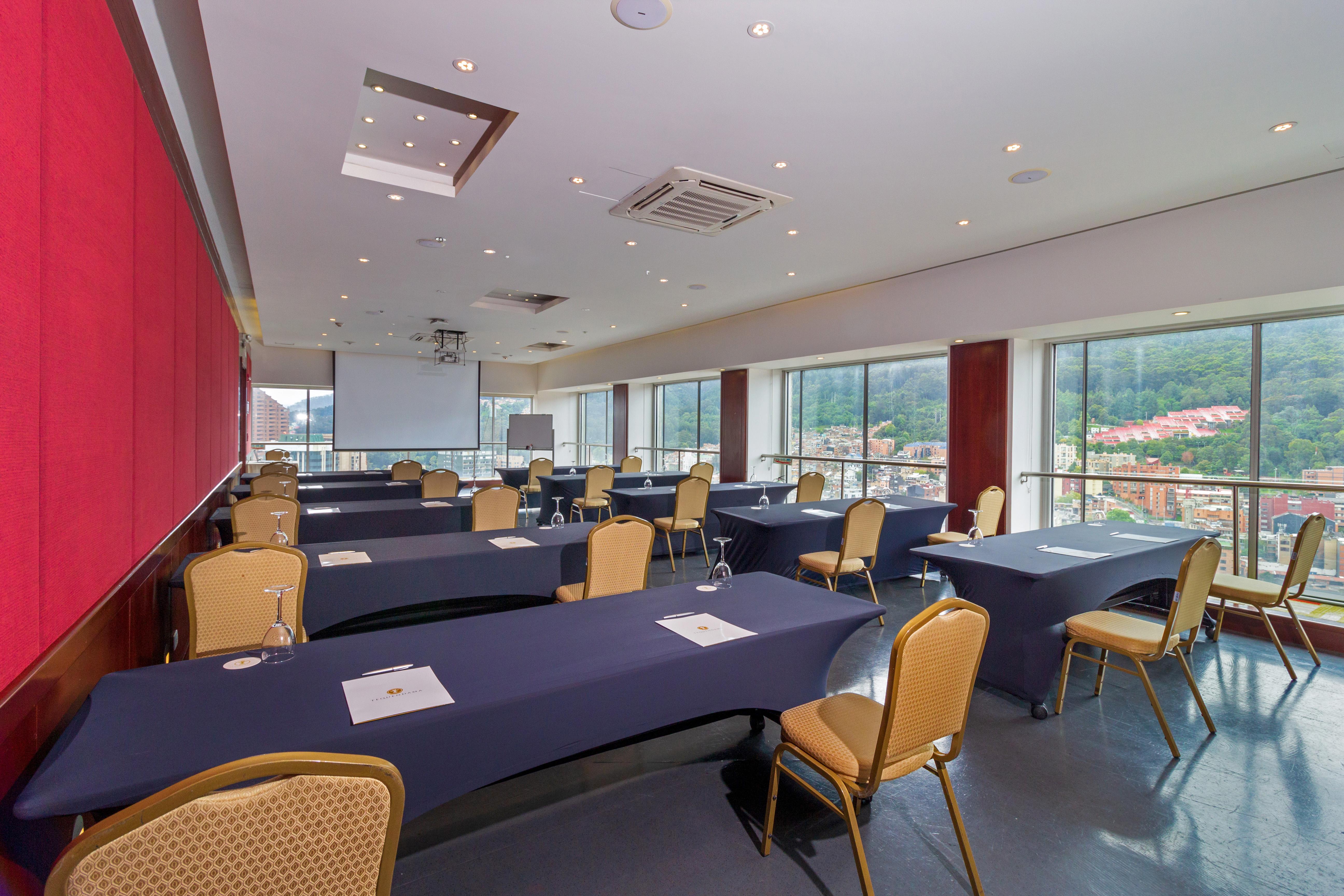 โรงแรมเตเกนดามา โบโกตา ภายนอก รูปภาพ The photo shows a conference room designed for meetings or presentations. It features several tables arranged in a rectangle, each covered with dark blue tablecloths. There are chairs with golden fabric surrounding the tables. The room has large wind