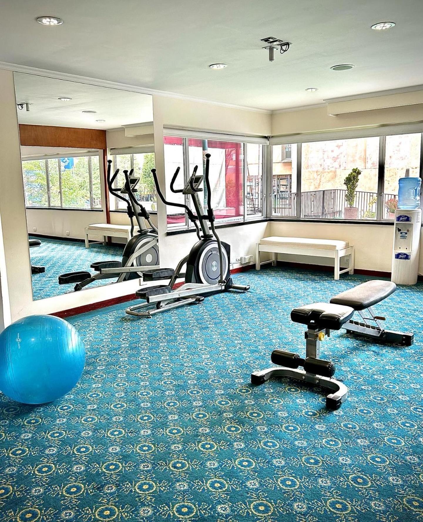โรงแรมเตเกนดามา โบโกตา ภายนอก รูปภาพ The photo shows a small gym or fitness area. In the space, there are two elliptical machines positioned next to large windows that allow natural light to enter. There's also a workout bench and a blue exercise ball on the floor, which features a patt