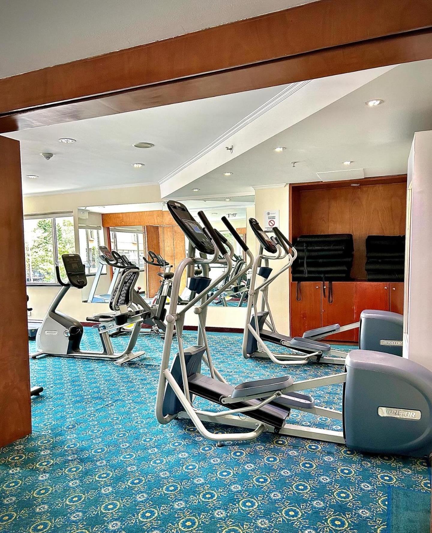 โรงแรมเตเกนดามา โบโกตา ภายนอก รูปภาพ The photo shows a fitness room equipped with several exercise machines. There are two elliptical machines and a couple of stationary bikes arranged in the space. The floor is covered with a blue carpet that has a patterned design. Large windows allow
