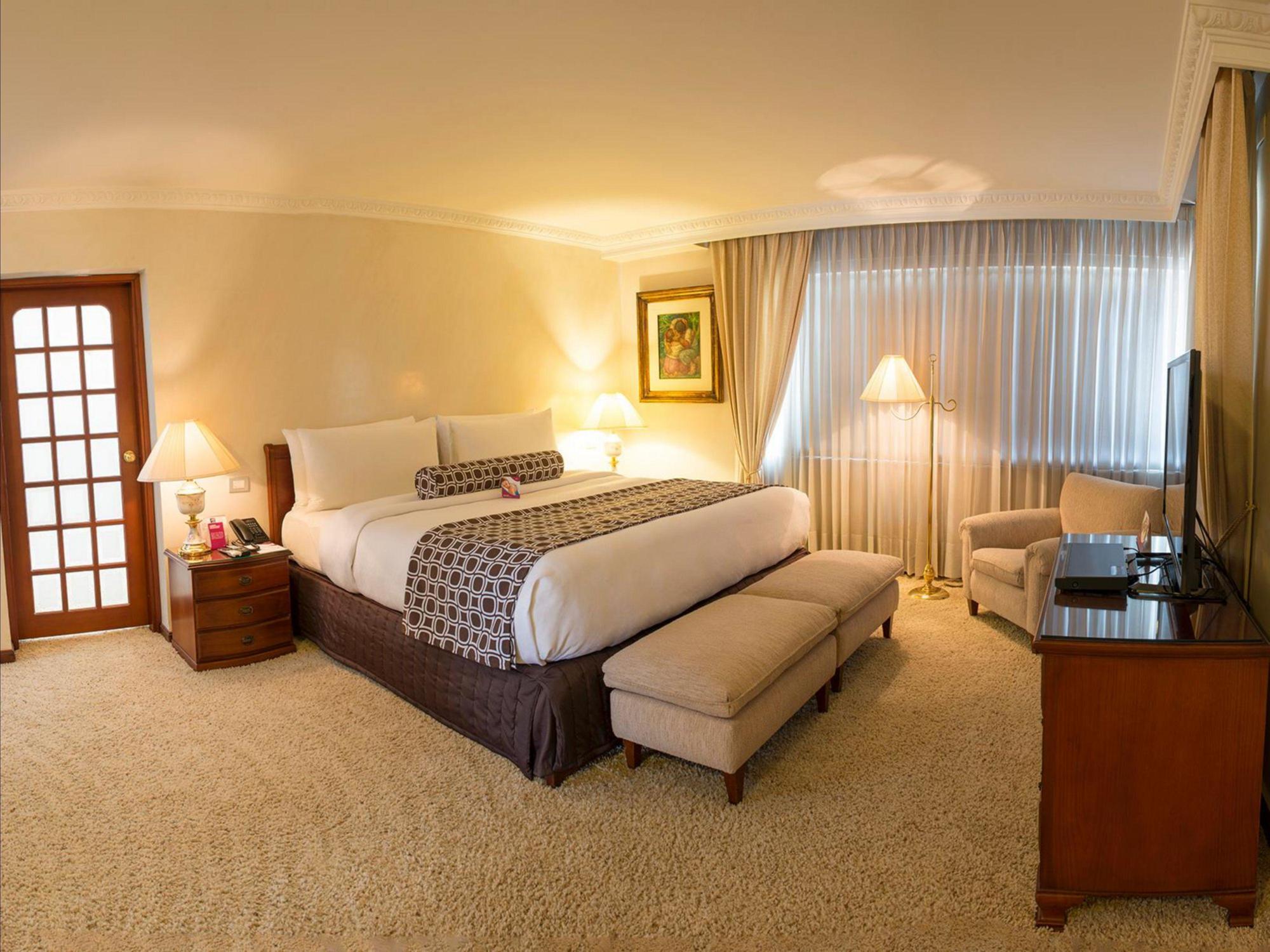 โรงแรมเตเกนดามา โบโกตา ภายนอก รูปภาพ The photo shows a well-appointed hotel room featuring a large bed with white bedding and a decorative throw pillow. There are two bedside lamps on wooden nightstands, adding a warm ambiance to the space. In front of the bed, there is a plush bench. O